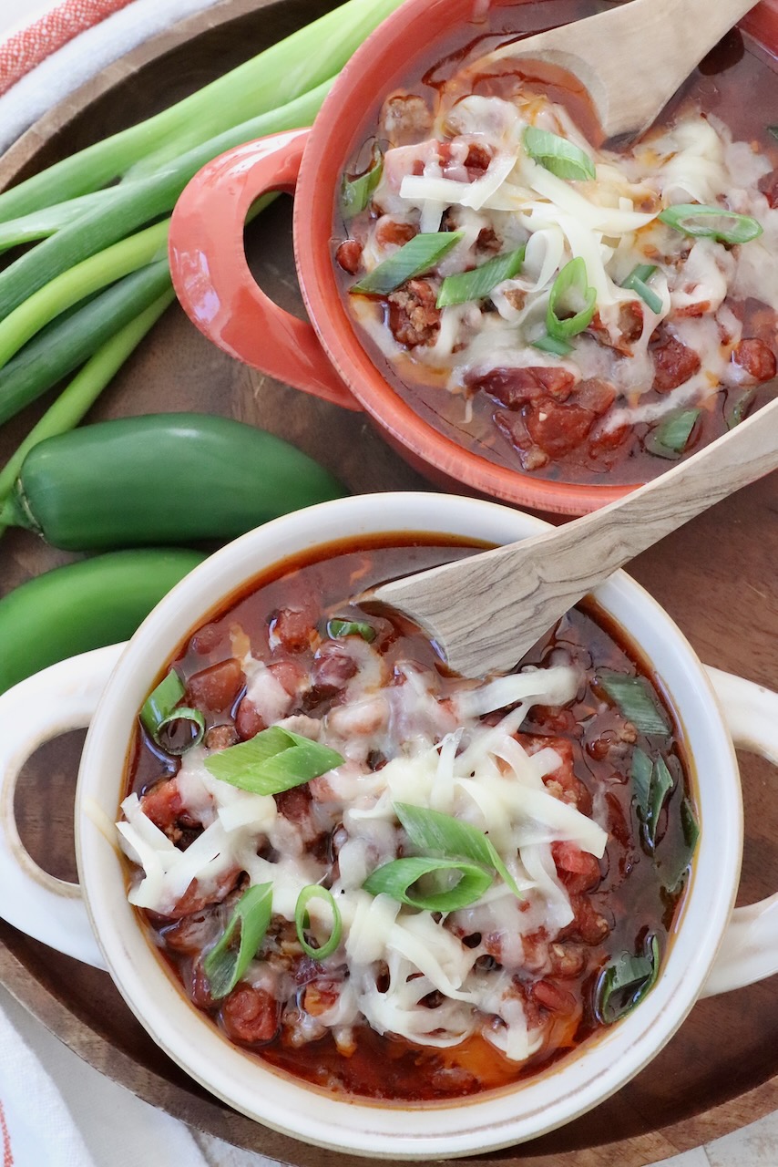 chili in bowls with spoons topped with shredded cheese and diced green onions