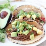Shredded beef tacos in corn tortillas on plate with avocado and cilantro