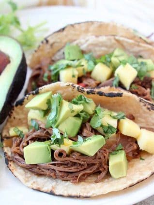 Shredded beef tacos in corn tortillas on plate with avocado and cilantro