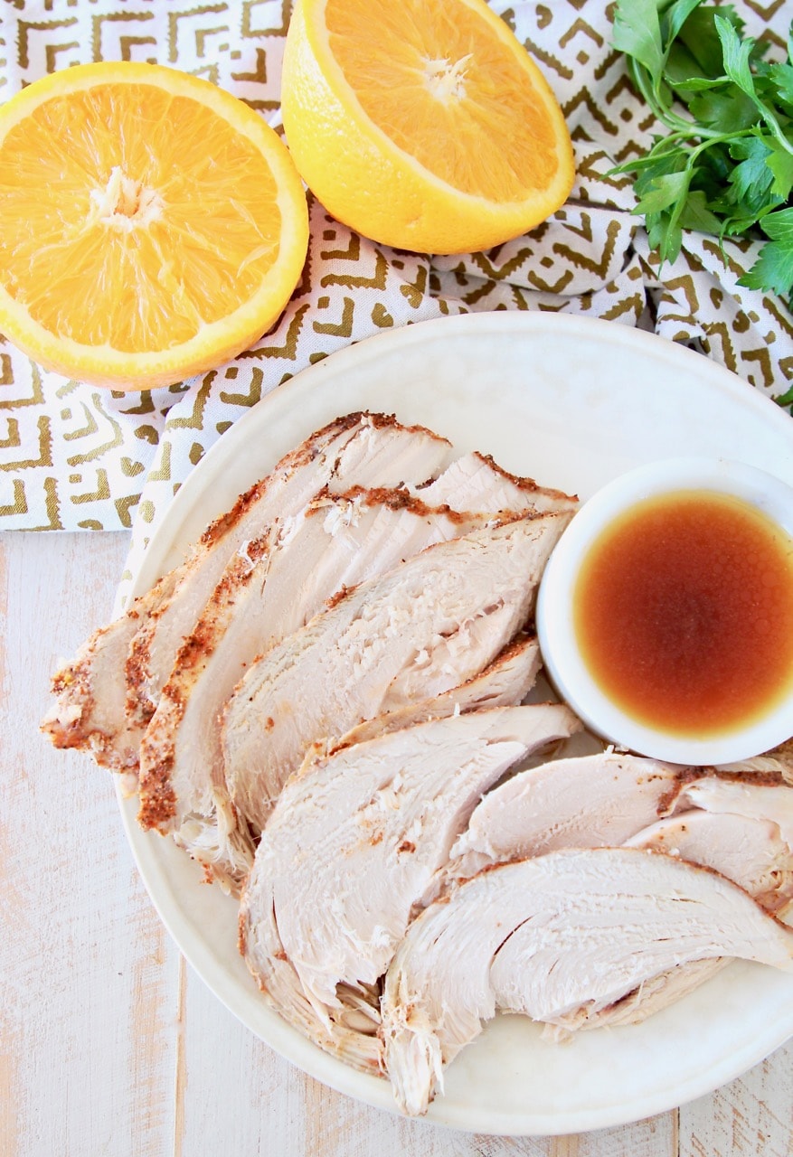 Slices of spice rubbed turkey breast on plate, next to small white bowl or orange bourbon sauce, with an orange cut in half next to the plate with fresh parsley