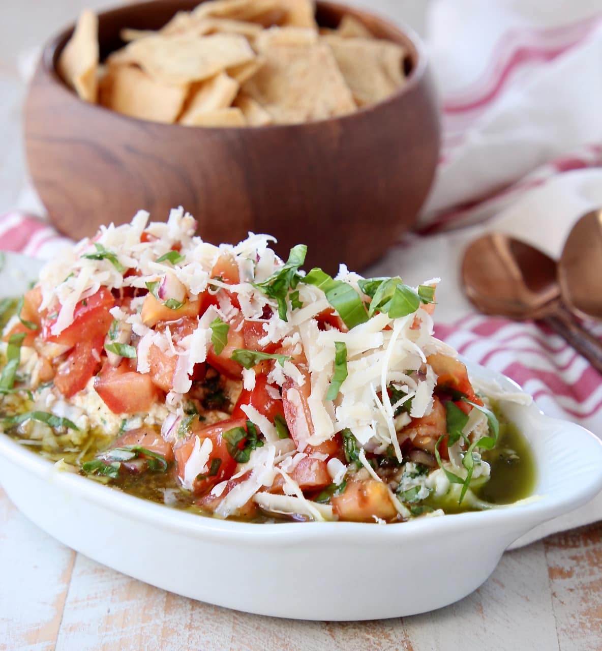 Italian 5 layer dip in white serving bowl with wooden bowl of pita chips behind it and 2 copper spoons next to it