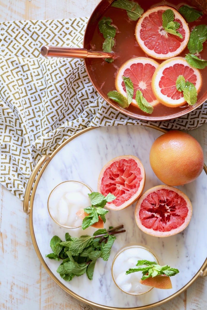 Rum punch in copper bowl with fresh grapefruit slices and mint springs, with a marble serving tray sitting next to it, topped with glasses of rum punch, grapefruits sliced open and fresh mint sprigs
