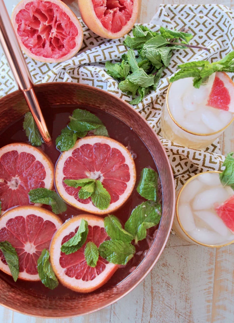 Copper serving bowl filled with spiced rum punch, topped with slices of grapefruit and mint leaves, with two glasses of rum punch on the side sitting on a patterned towel with fresh mint leaves