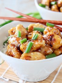 Pieces of Chinese Honey Chicken in white bowl with white rice, garnished with chopped green onions and sesame seeds with brown chopsticks sitting on the side of the bowl