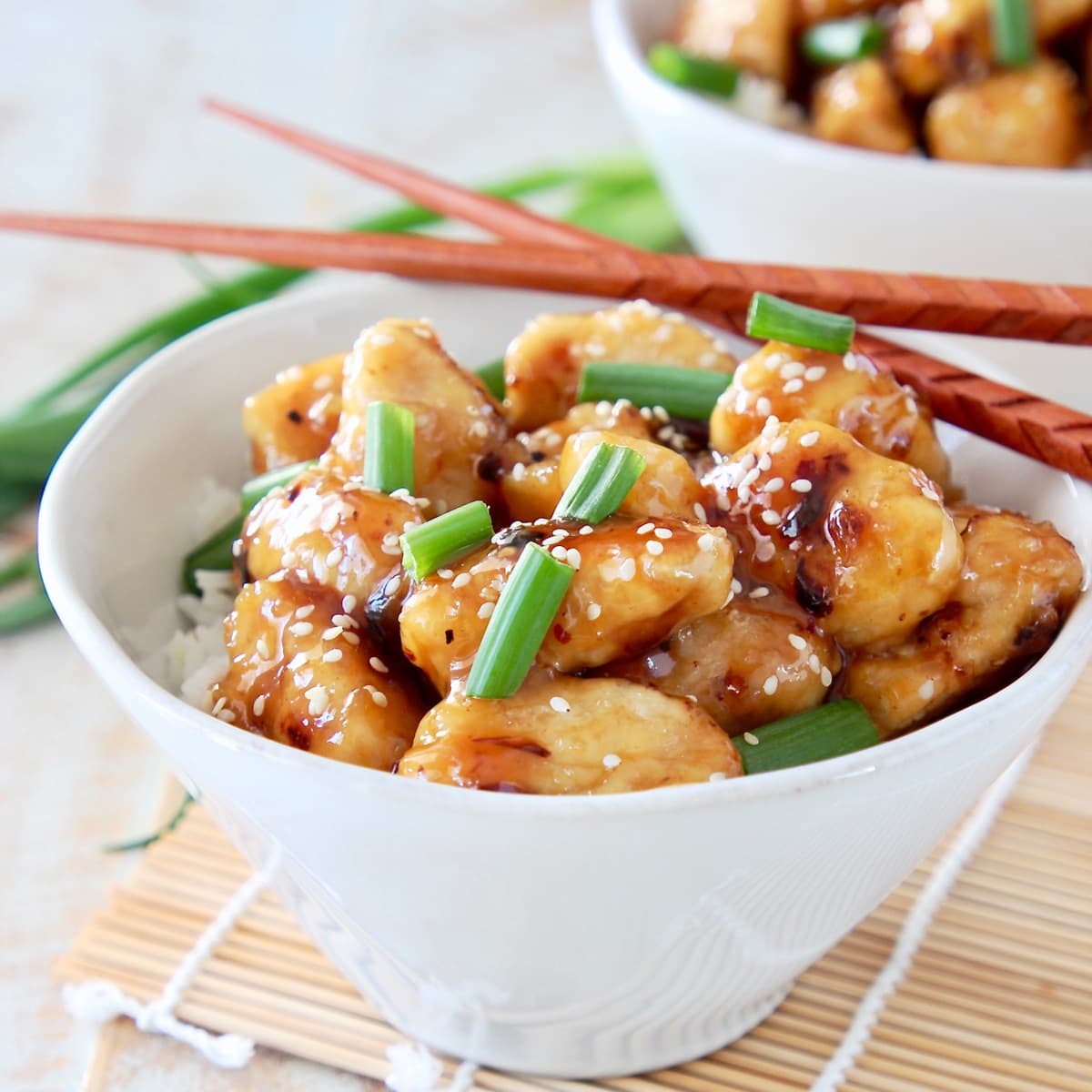 Pieces of Chinese Honey Chicken in white bowl with white rice, garnished with chopped green onions and sesame seeds with brown chopsticks sitting on the side of the bowl