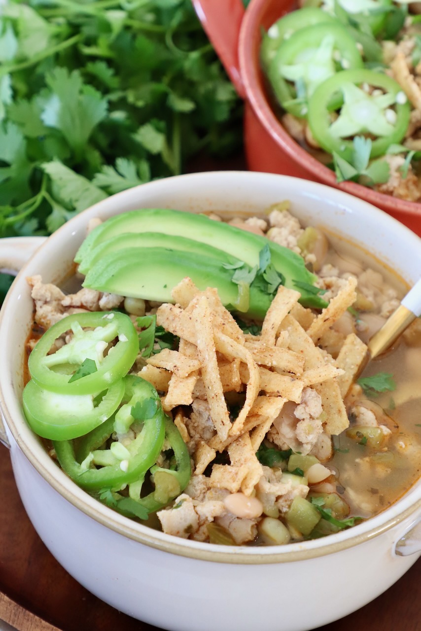 white chicken chili in bowl topped with sliced jalapeno, avocado and tortilla strips
