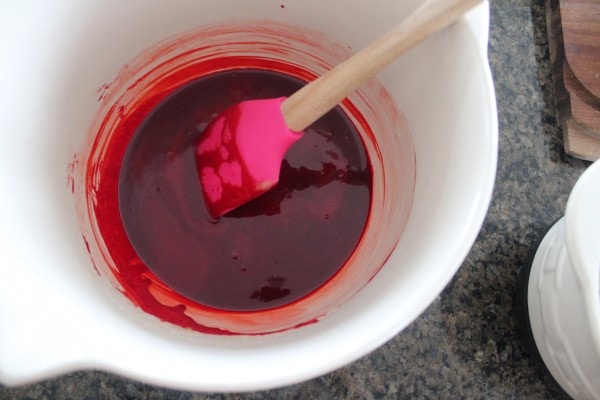 Butter, sugar, vanilla, eggs, and red food coloring mixed in a white bowl. 