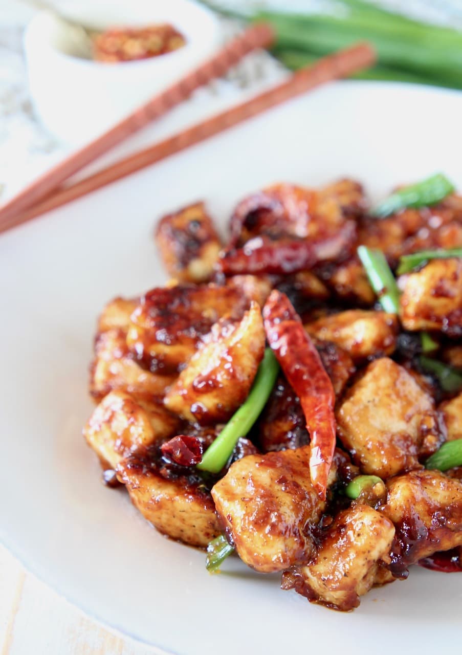 Gluten free general tso chicken with red chili peppers and green onions on white plate with wood chopsticks