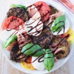 A vibrant Caprese salad with sliced tomatoes, mozzarella, crispy eggplant, prosciutto, fresh basil, and balsamic glaze drizzle on a white plate.