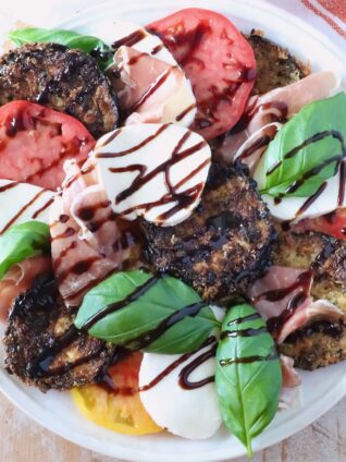 A vibrant Caprese salad with sliced tomatoes, mozzarella, crispy eggplant, prosciutto, fresh basil, and balsamic glaze drizzle on a white plate.