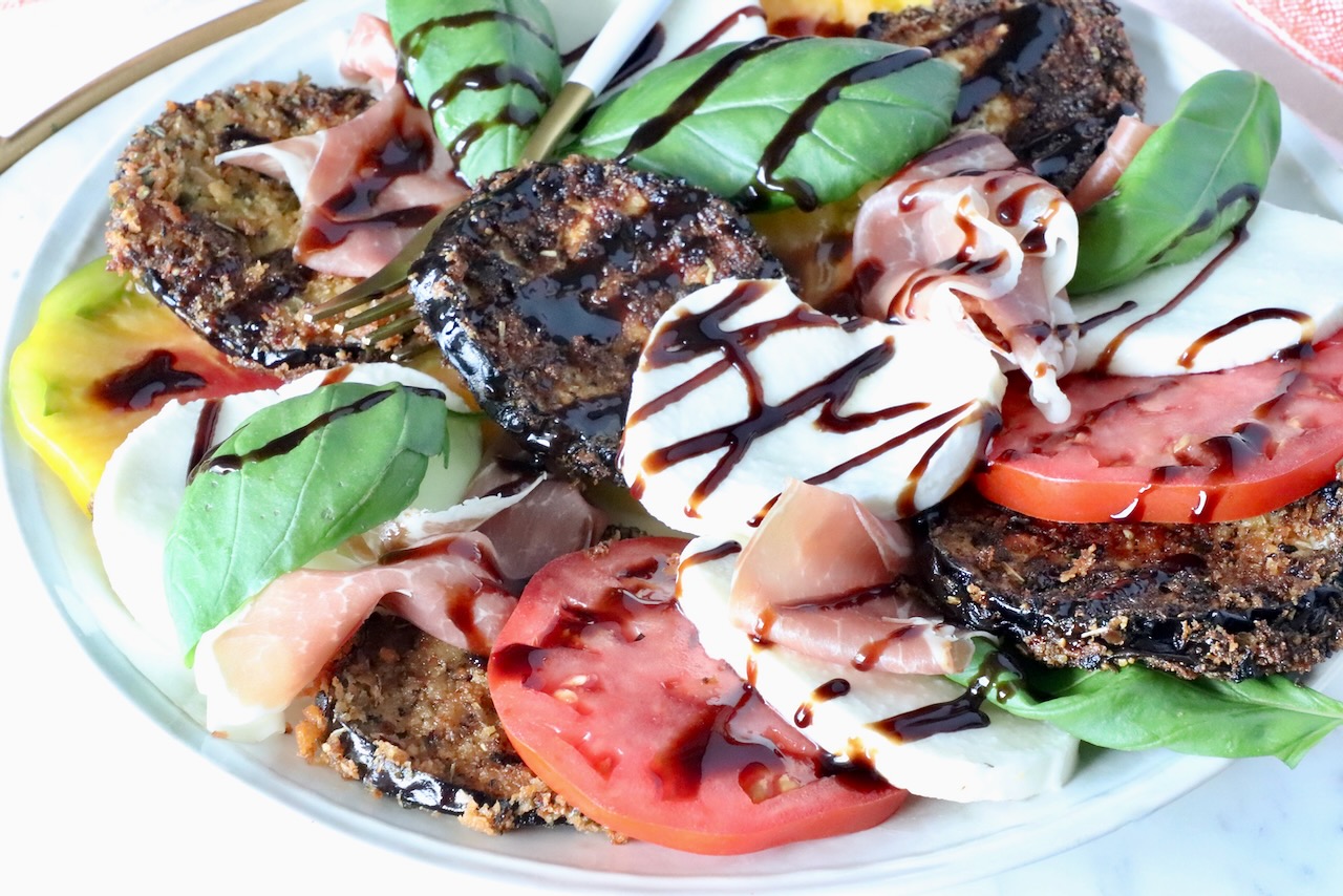 A colorful salad featuring breaded eggplant slices, fresh mozzarella, ripe tomatoes, basil leaves, and prosciutto, drizzled with balsamic glaze.