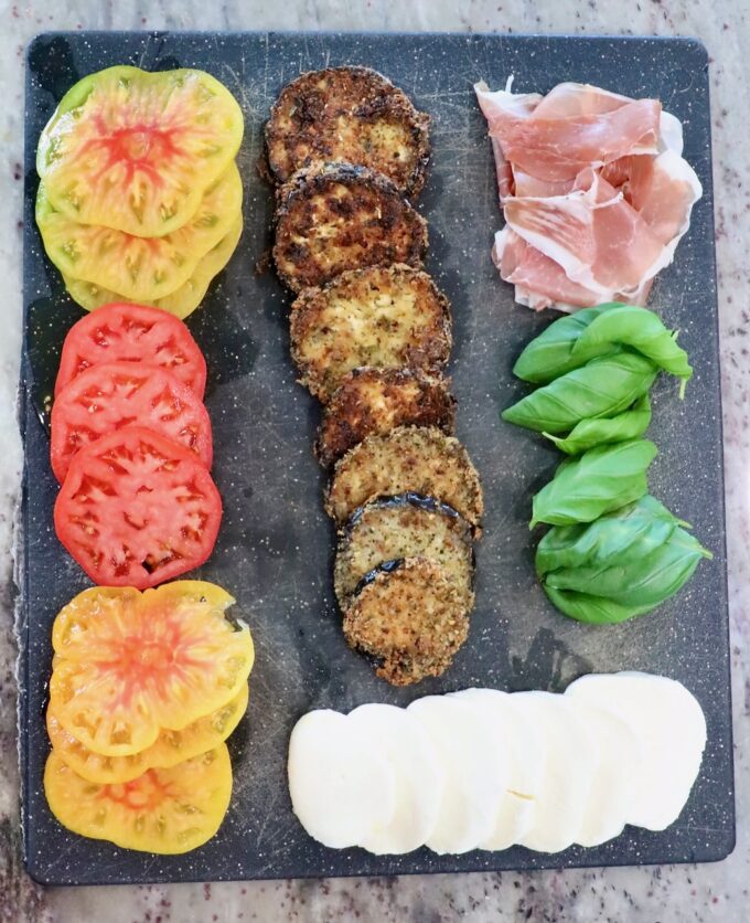 sliced heirloom tomatoes, crispy fried eggplant slices, sliced mozzarella cheese, basil leaves, and slices of prosciutto on a black cutting board