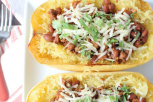 Turkey chili inside of roasted spaghetti squash halves on white plate with shredded cheese and cilantro on top with two orange forks on the side