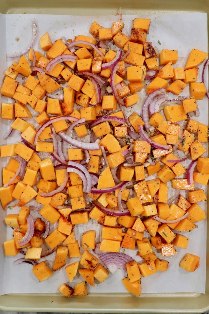 cubed butternut squash with sliced onions and minced garlic on a parchment paper lined baking sheet