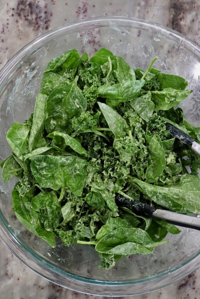 baby spinach and kale tossed in a creamy dressing in a large glass bowl with tongs