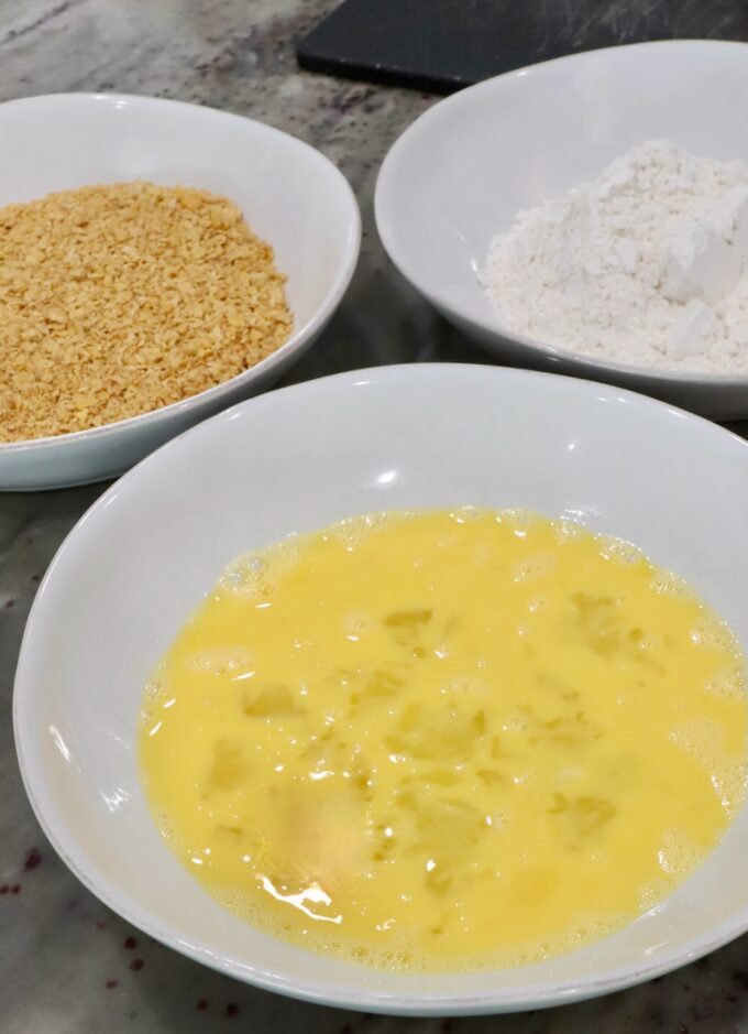 three bowls on a countertop, one with whisked eggs, one with flour, and one with crushed cornflakes