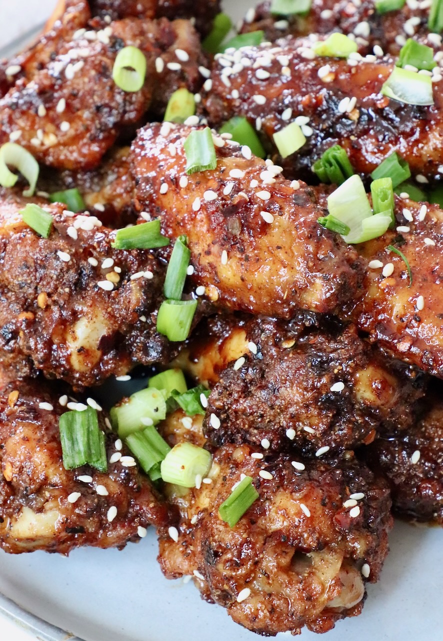 cooked chicken wings tossed in sauce piled up on a plate, topped with diced green onions and sesame seeds