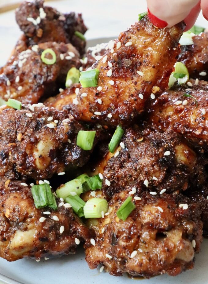 hand grabbing a chicken wing tossed in sauce piled up on other cooked chicken wings on a plate