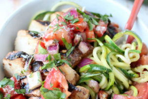 Zucchini noodles wrapped around fork in bowl with grilled chicken and fresh tomato bruschetta