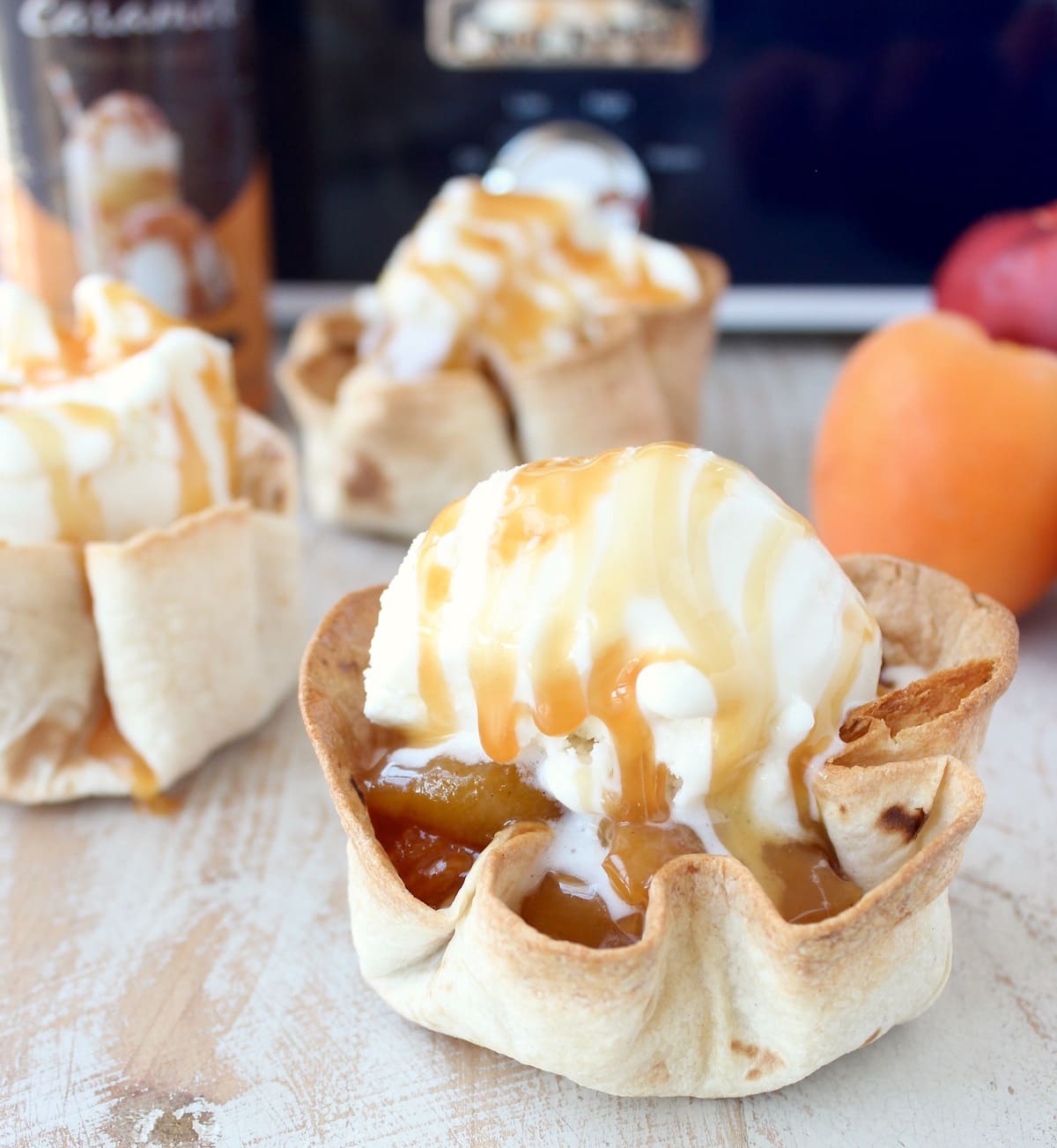 crispy taco shell cups filled with slow cooked fruit, topped with a scoop of ice cream and caramel syrup on a white wood board in front of a blue crock pot