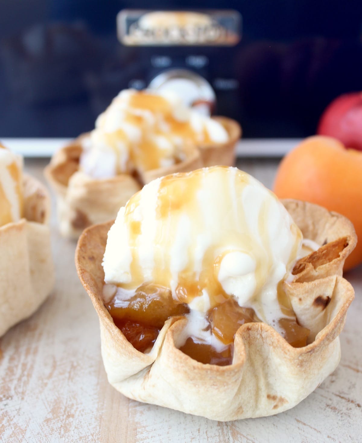 crispy taco shell cups filled with slow cooked fruit, topped with a scoop of ice cream and caramel syrup on a white wood board in front of a blue crock pot