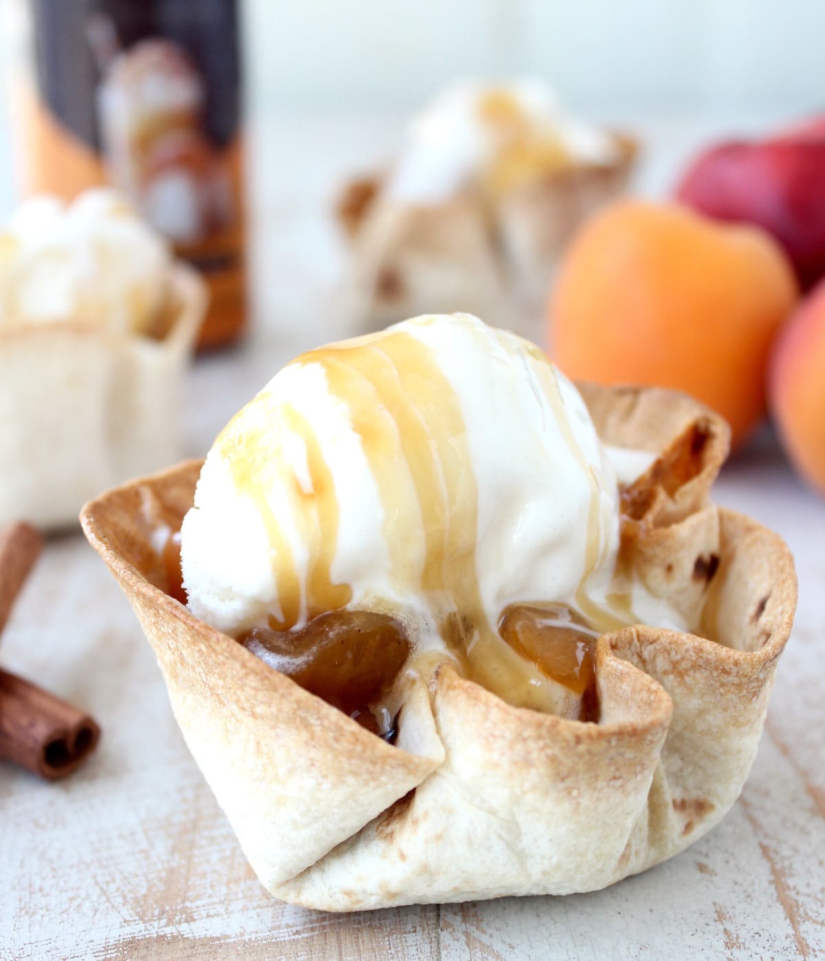 crispy taco shell cups filled with cooked fruit, topped with a scoop of ice cream and caramel syrup on a white wood board