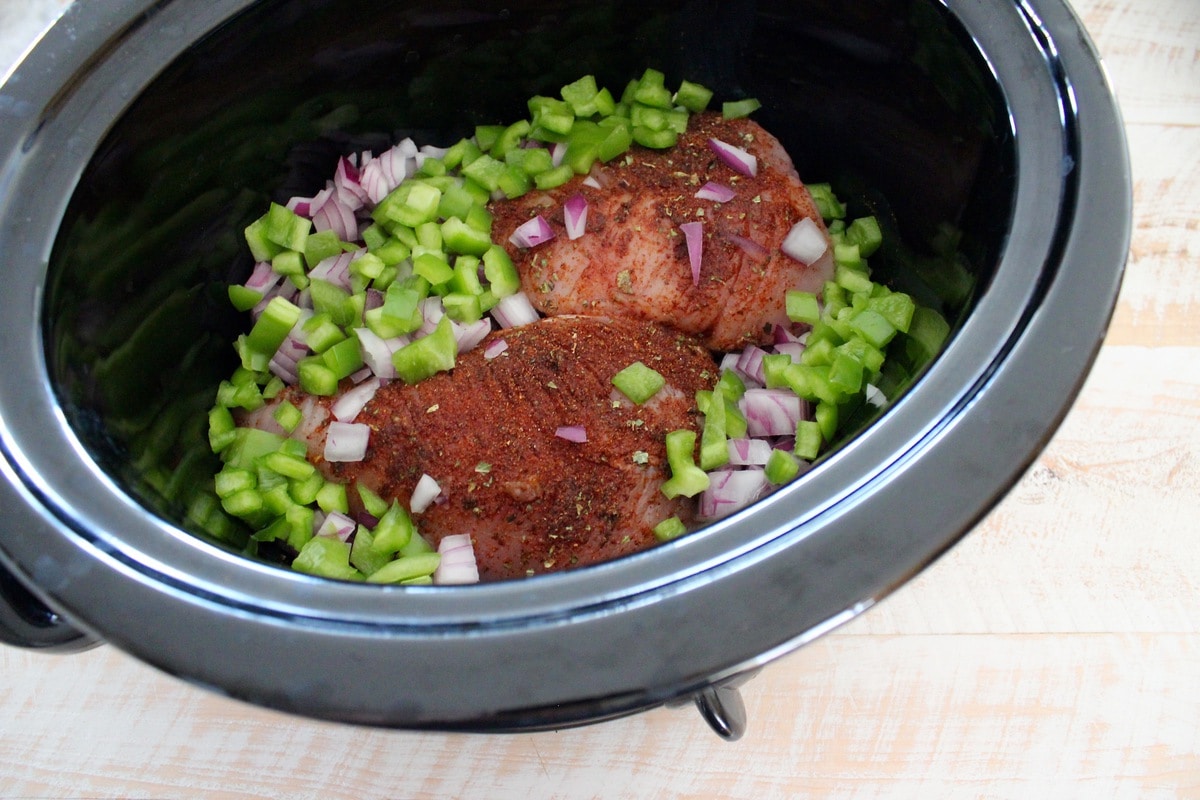 A black crock pot bowl filled with seasoned chicken, diced peppers, and onions.