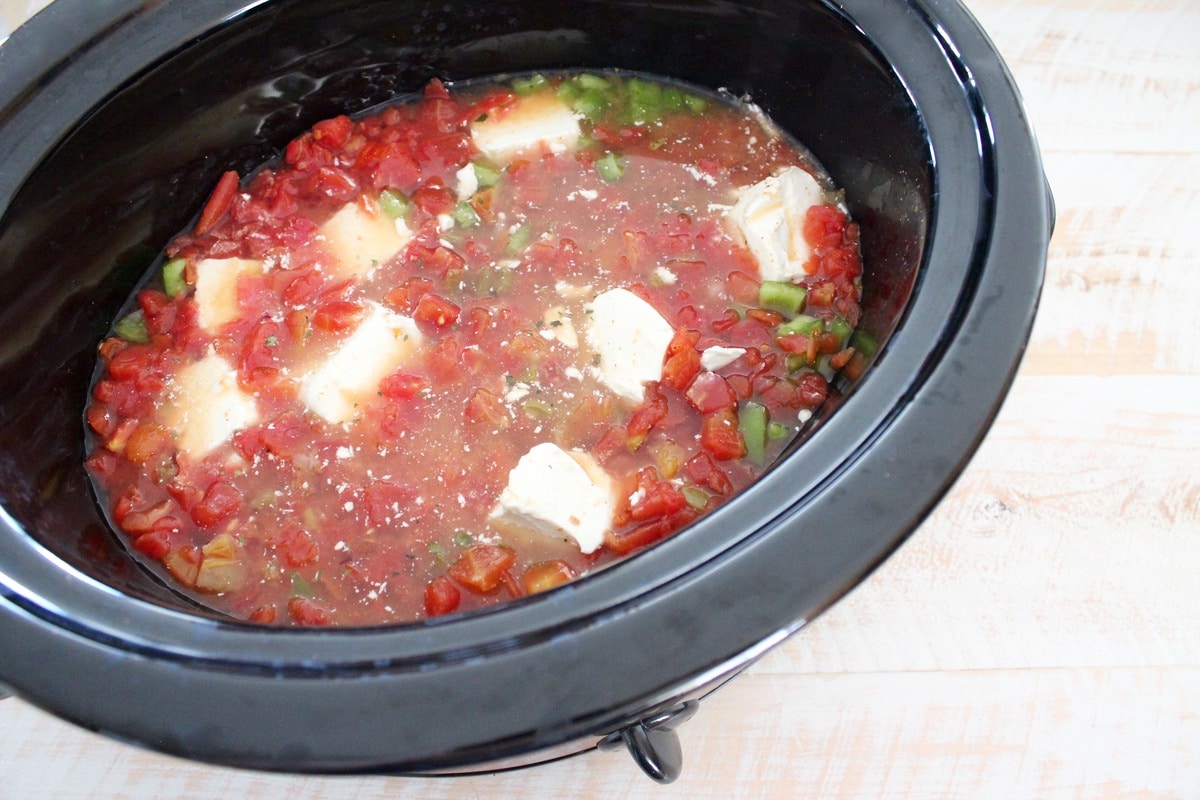 A slow cooker filled with diced peppers, diced tomatoes, broth, and chunks of cream cheese.