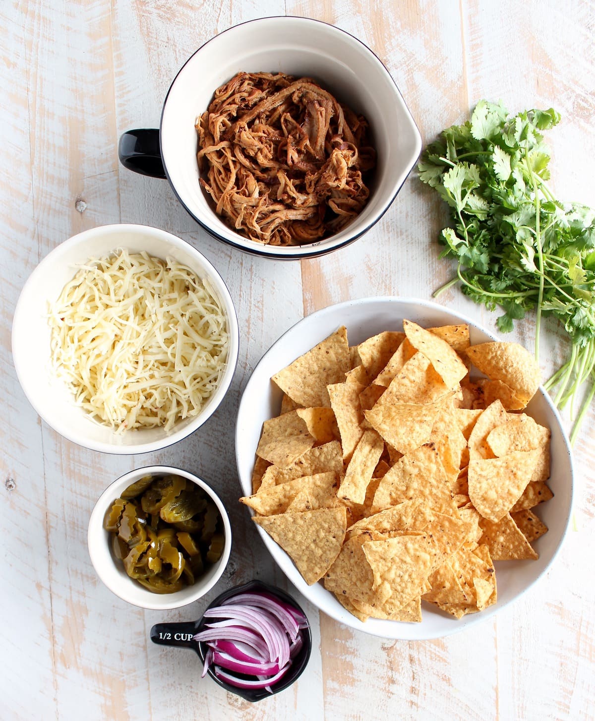 The Ultimate Pulled Pork Nachos Recipe Ingredients in bowls; chips, sliced red onions, jalapeños, cheese, pulled pork, and cilantro 