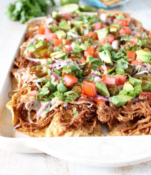 A white dish filled with tortilla chips topped with pulled pork, cheese, onions, tomatoes, and avocado.