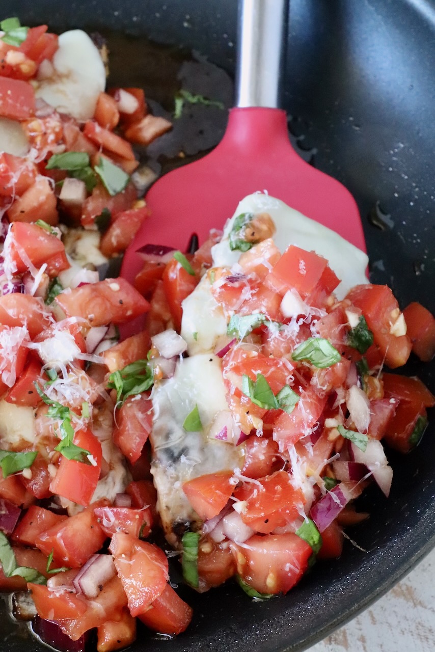 cooked chicken topped with cheese and tomato bruschetta in a skillet with a red spatula