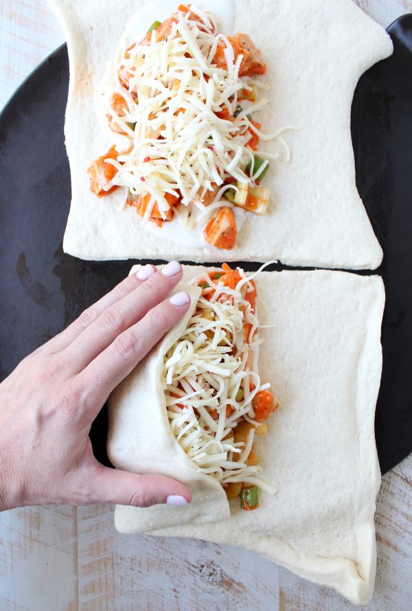 Hand folding uncooked pizza dough over chicken, celery, carrots, green onions, buffalo sauce and cheese.