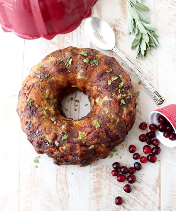 Sausage stuffing that has been cooked in a bundt pan surrounded by a spoon, herbs, and cranberries.