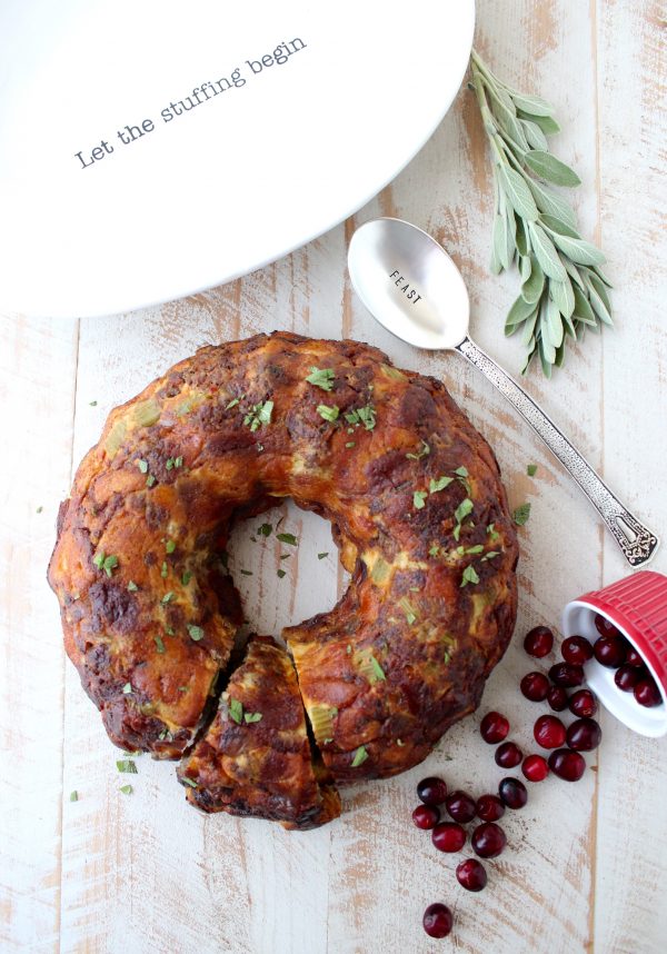 Stuffing that has been cooked in a bundt pan on a wooden surface surrounded by a spoon, herbs, and cranberries.