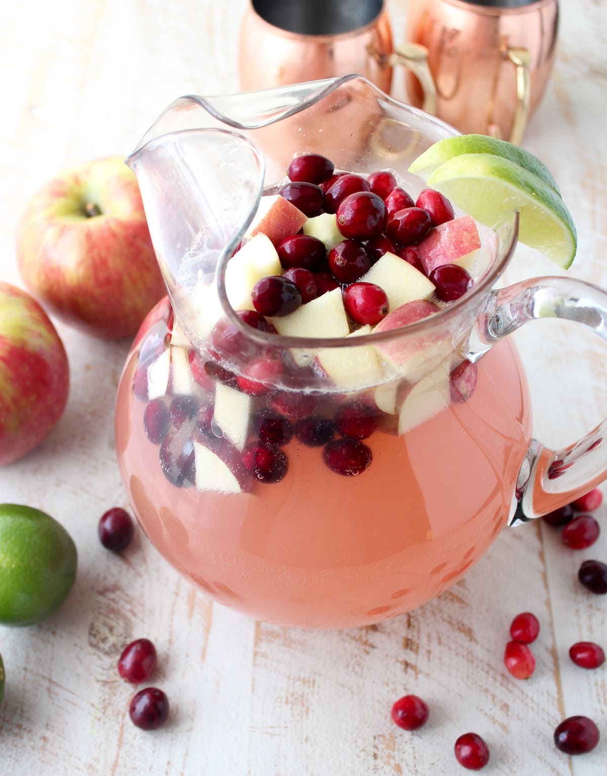 cranberry apple moscow mule punch in a large glass pitcher with fresh cranerries and diced apples