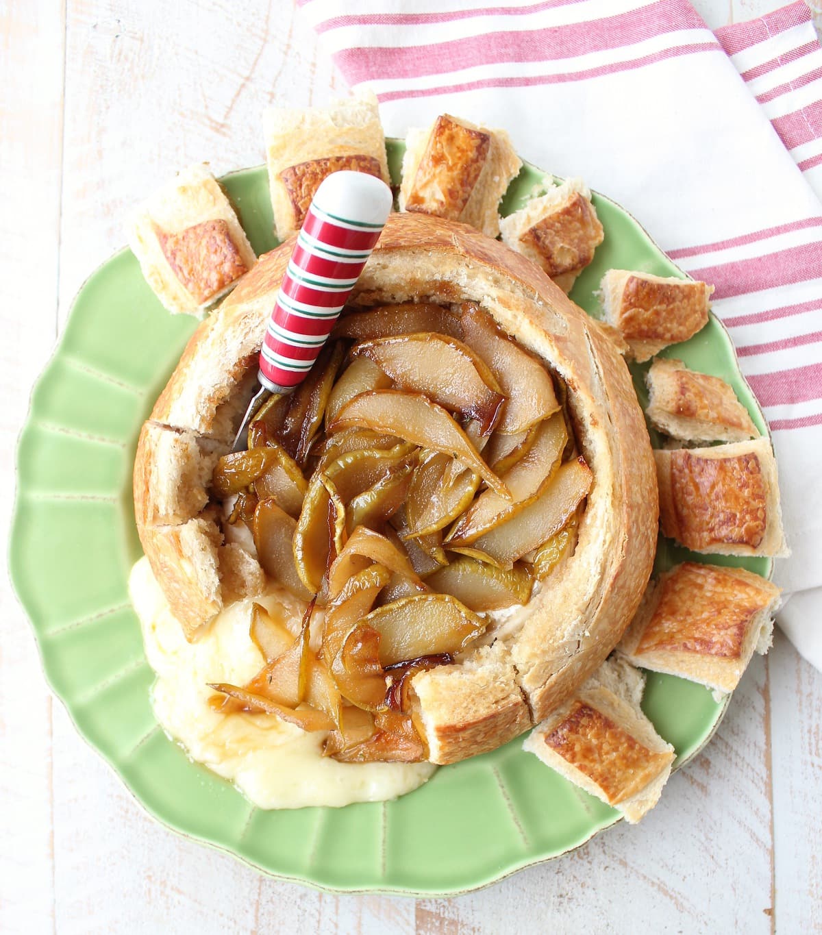 Baked brie in bread bowl with caramelized pears and honey on green plate with red and white cheese spreader and towel