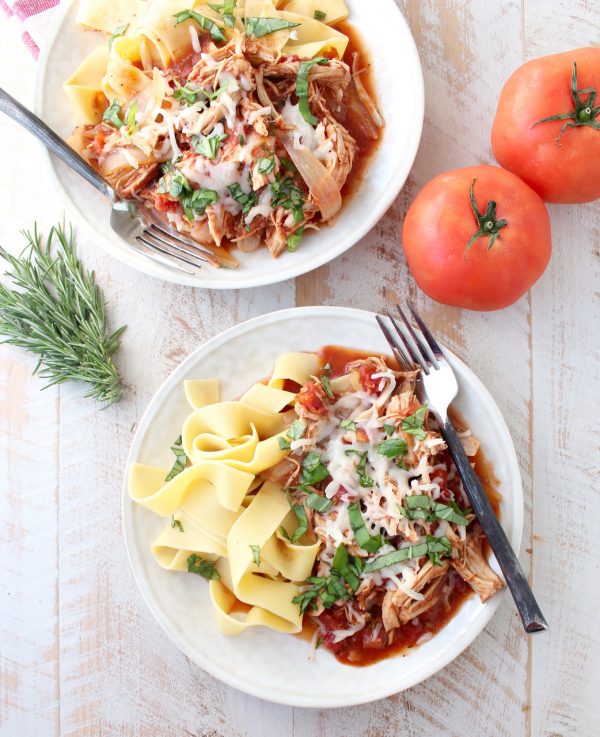 Two white plated topped with pasta, slow cooker chicken ragu, melted cheese, and fresh herbs all on a wooden surface.