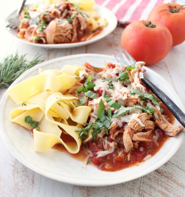 Shredded chicken covered in red sauce, cheese, and herbs with pasta on a white plate.