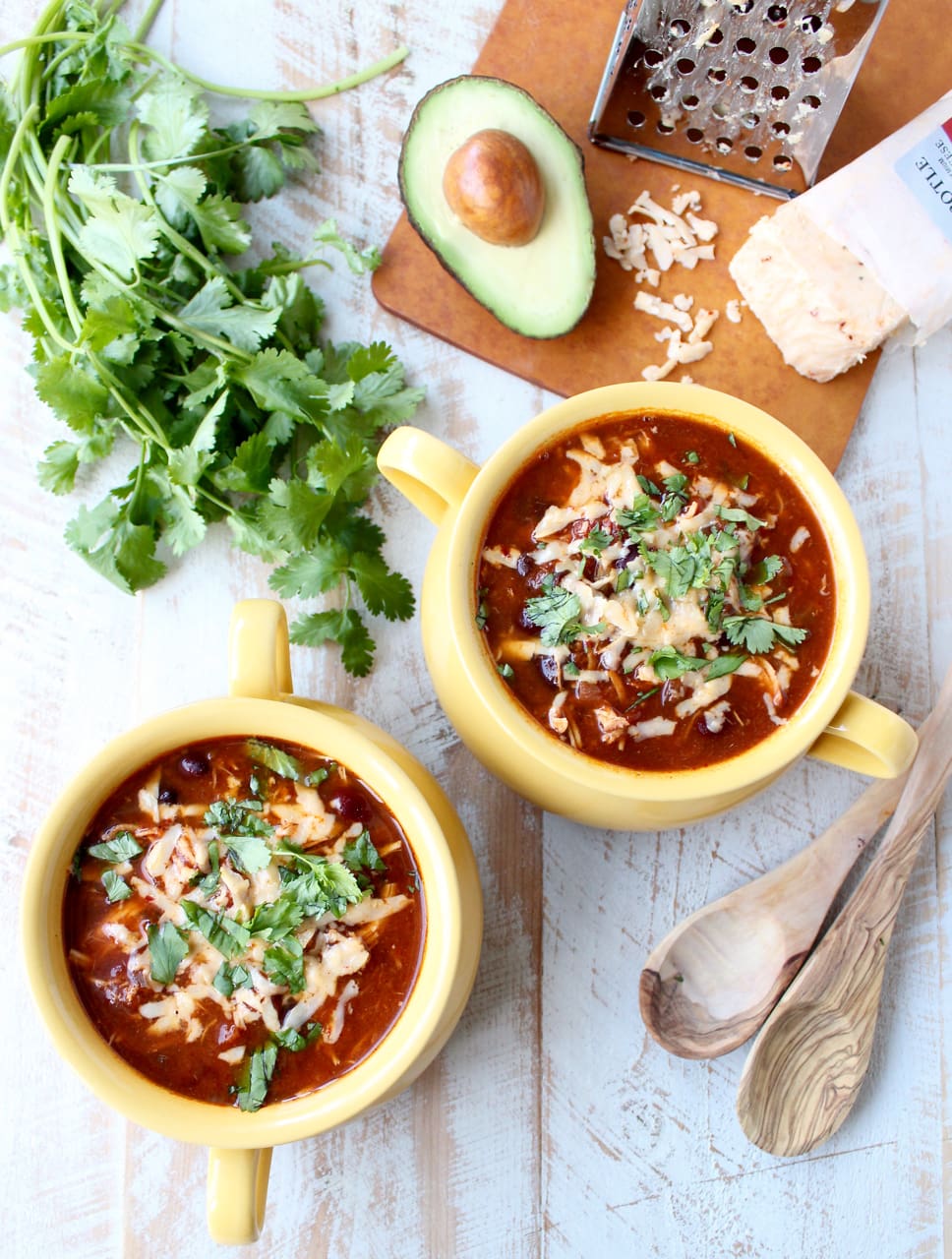 Two yellow bowls on a wooden surface filled with enchilada soup, cheese, and cilantro.