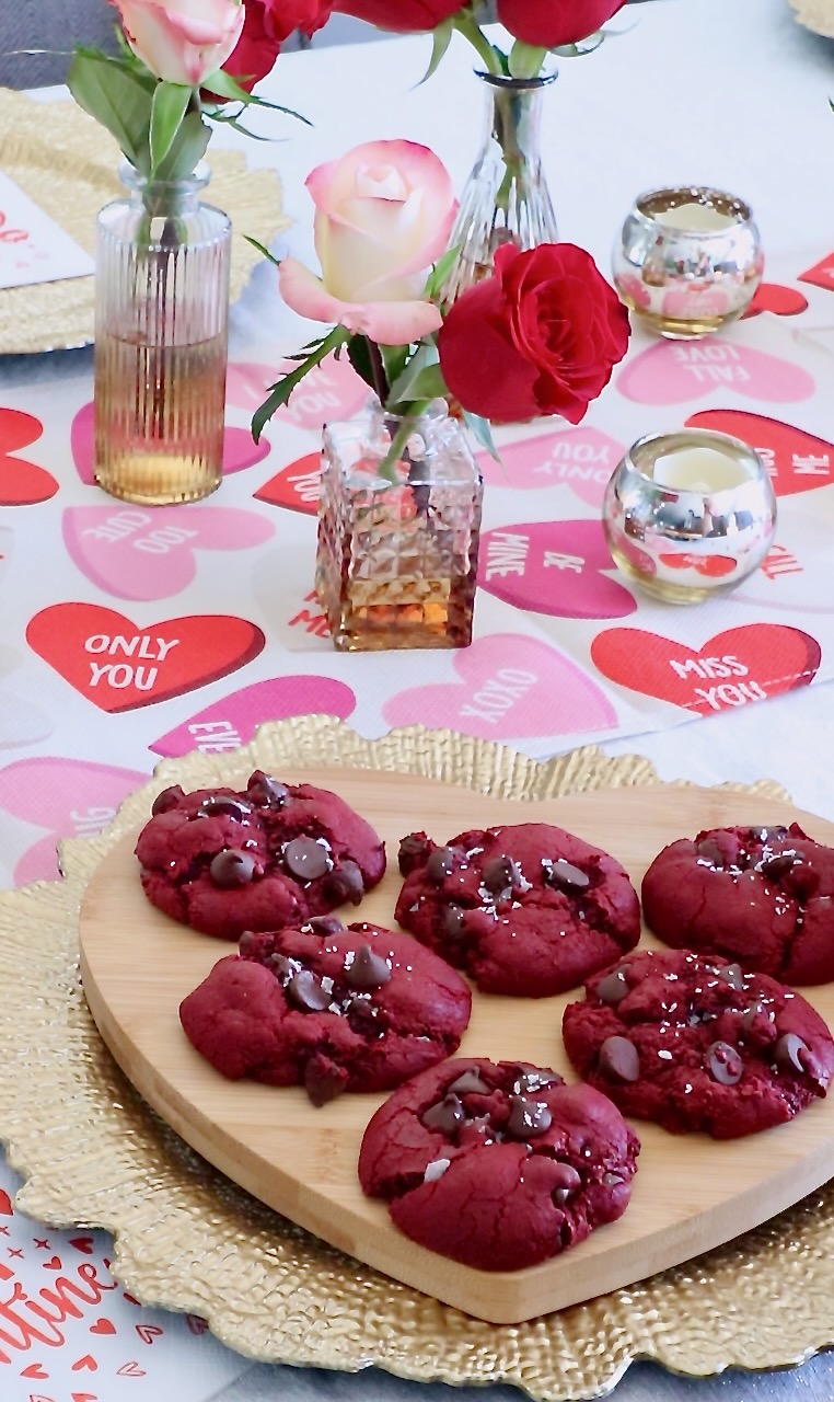 Heart-shaped wooden tray with red velvet cookies, chocolate chips on top. Surrounding are roses in vases and heart-themed table decor.