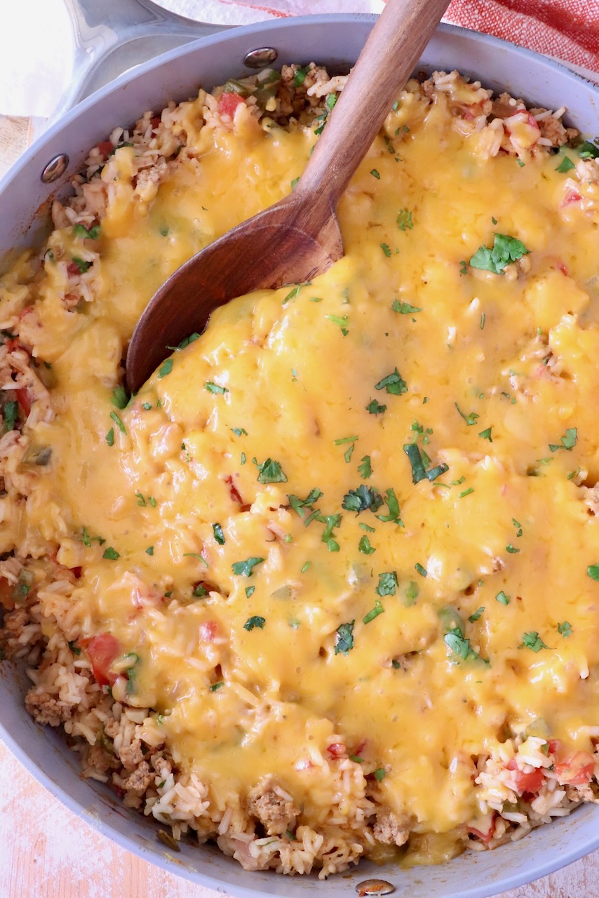 A skillet filled with cheesy Mexican rice, topped with melted golden cheese and garnish of green herbs. A wooden spoon is scooping from the side.