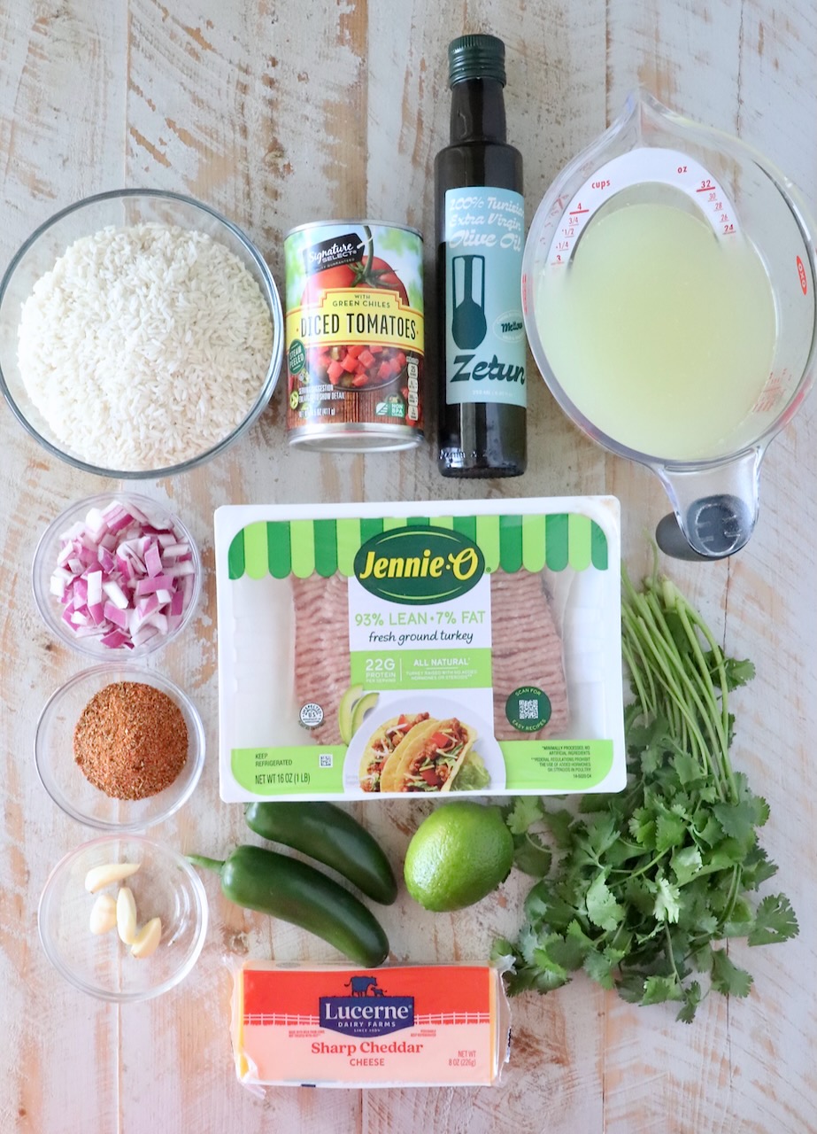 Ingredients for a recipe displayed on a wooden surface include ground turkey, cheddar cheese, diced tomatoes, rice, chicken broth, olive oil, cilantro, lime, jalape&ntilde;os, garlic, chopped red onion, and a spice mix.