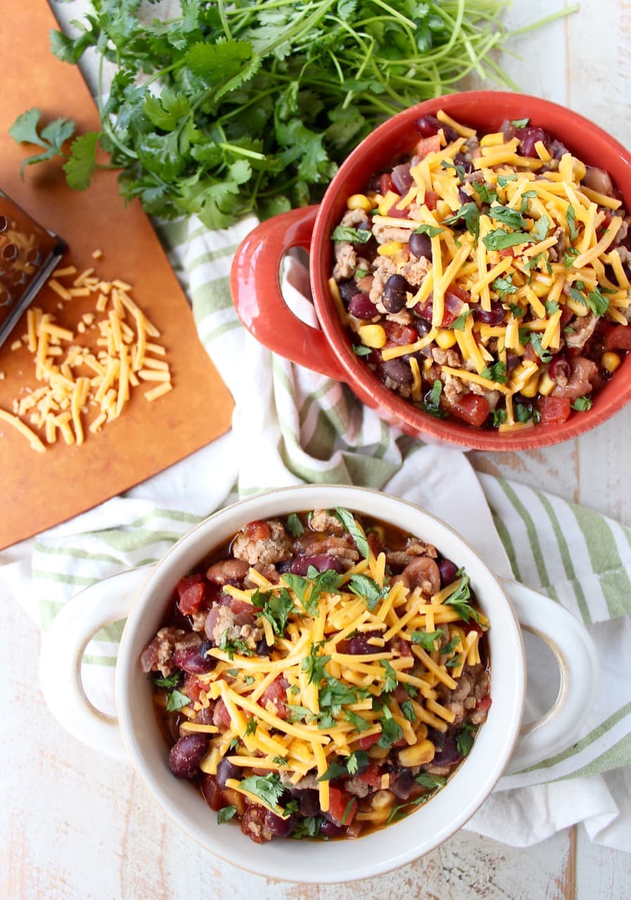 Two bowls filled with chili and shredded cheese on a wooden surface surrounded by a green and white napkin, spoons, herbs, and a cutting board with shredded cheese.