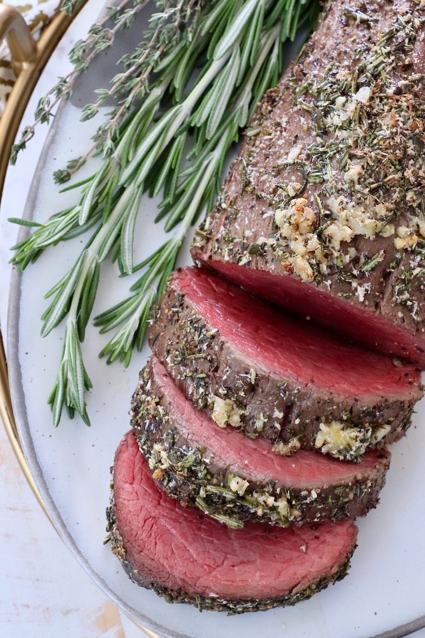 herb crusted beef tenderloin partially sliced on a plate with fresh herbs