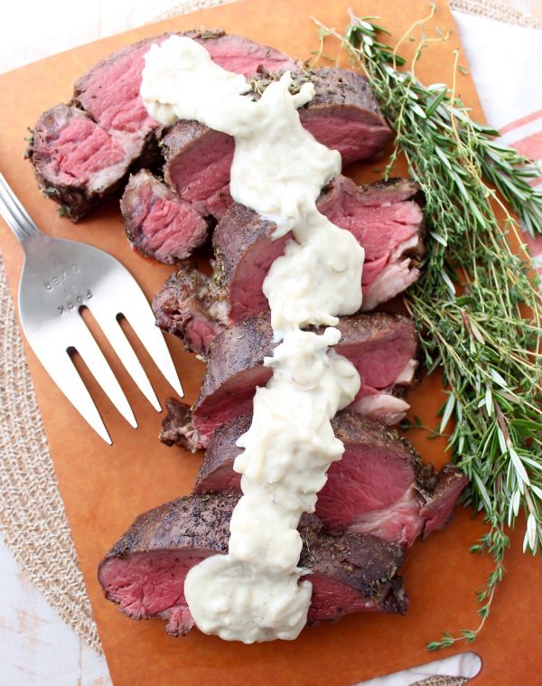 Sliced beef tenderloin topped with a creamy sauce on a cutting board aside herbs. 