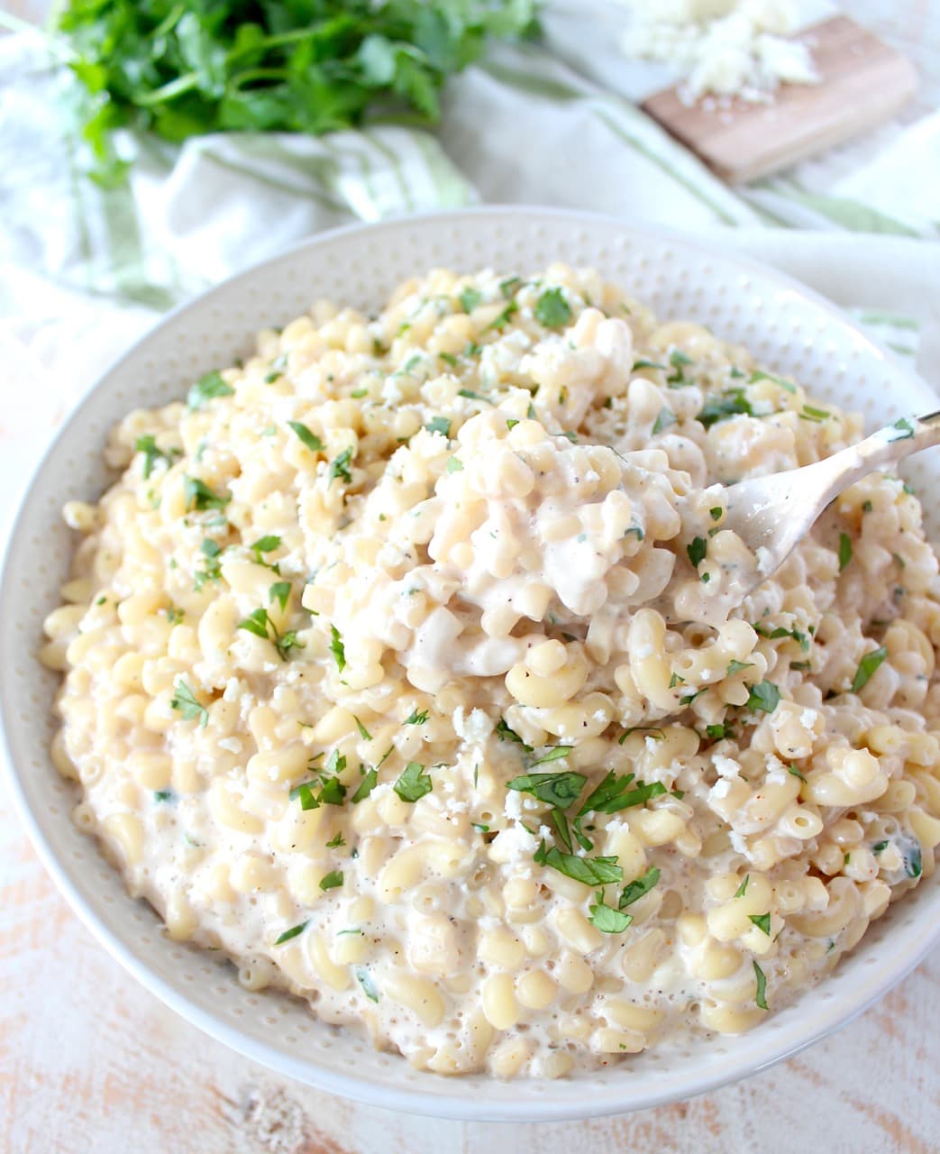 Mexican street corn mac and cheese in a white bowl with green herbs in the background. 