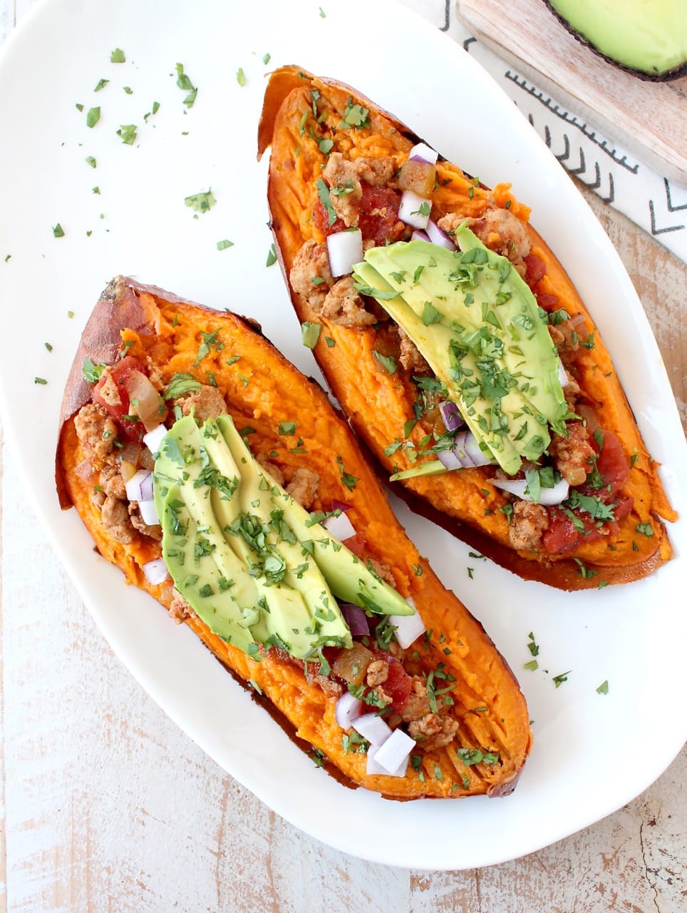 baked sweet potato sliced in half on white plate, topped with chili, avocado and cilantro