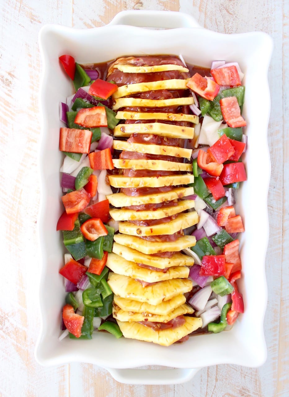 uncooked pork tenderloin filled with slices of pineapple in a baking dish surrounded by diced bell peppers and onions