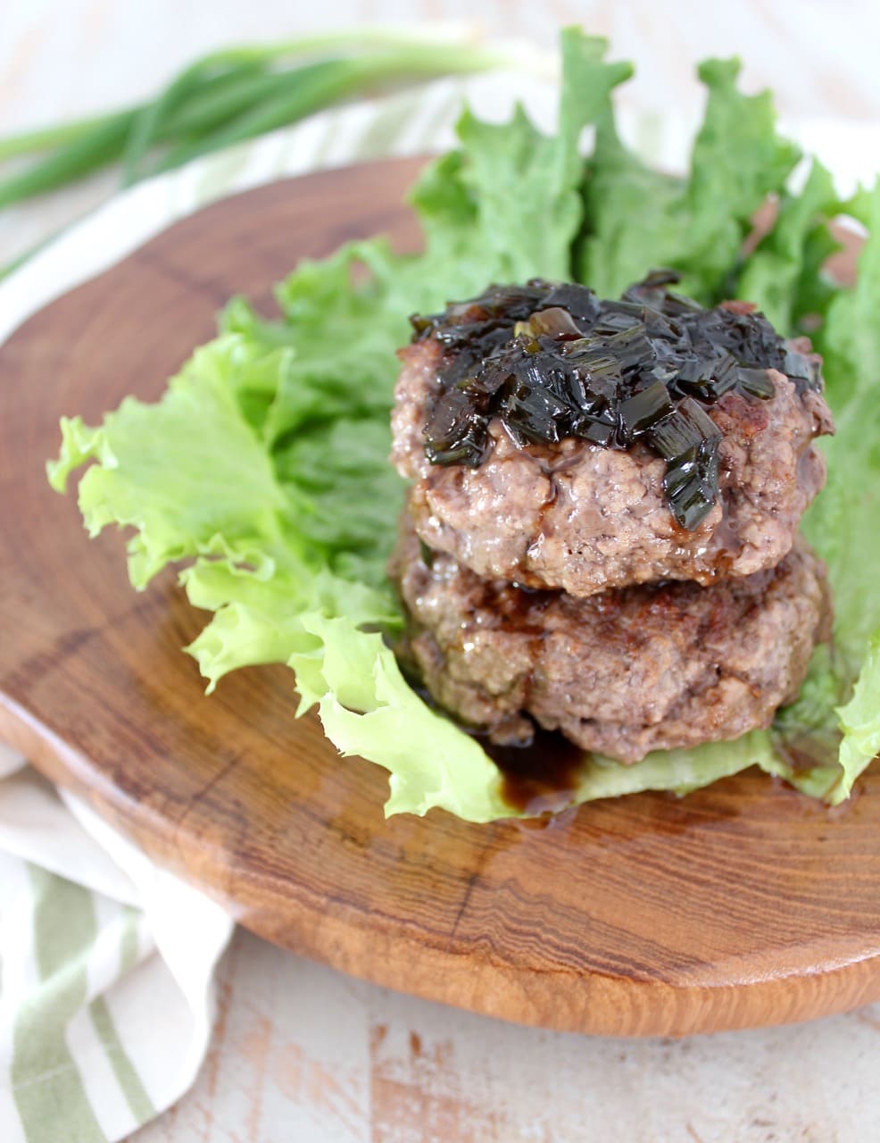 two cooked burger patties stacked on top of each other on a leaf of lettuce, topped with a scallion sauce