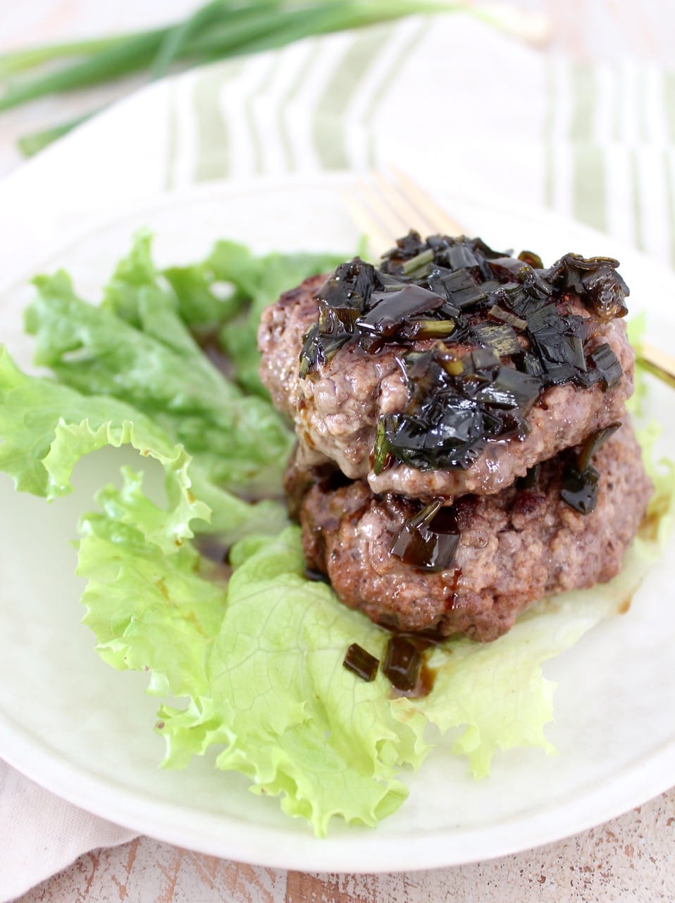 two cooked burger patties stacked on top of each other on a leaf of lettuce, topped with a scallion sauce